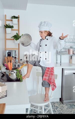 Vue latérale pleine longueur d'enfant pieds nus émerveillé dans le chapeau de chef et la veste ouvrant la casserole tout en cuisinant sur la cuisinière et debout sur la chaise dans la cuisine à la maison Banque D'Images