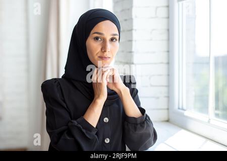 Portrait d'une jeune femme musulmane dans un hijab noir debout à l'intérieur près de la fenêtre, pliant ses bras et posant et regardant sérieusement la caméra. Banque D'Images