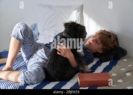 Garçon serein aux cheveux bouclés se détendant sur le lit tout en caressant chiot moelleux dans la chambre claire à la maison Banque D'Images