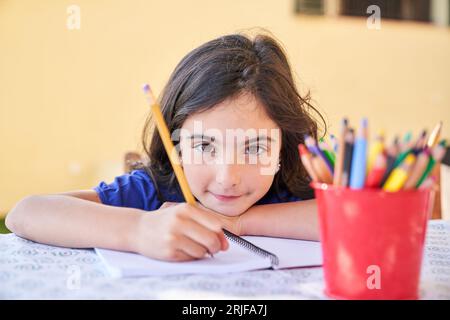 Inspiré petit enfant de l'école primaire utilisant un crayon pour écrire sur du papier blanc et assis au bureau tout en regardant la caméra Banque D'Images