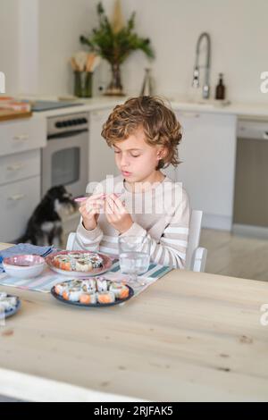 Garçon concentré dans des vêtements décontractés assis à une table en bois et mangeant de délicieux rouleaux de sushi avec des baguettes roses dans la cuisine à la maison Banque D'Images