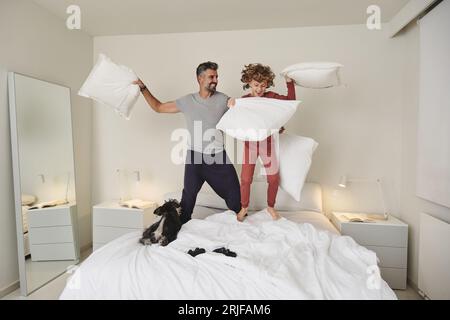 Corps entier de père barbu riant et enfant bouclé pieds nus en pyjama sautant sur le lit avec le chien près du miroir tout en se battant avec des oreillers Banque D'Images