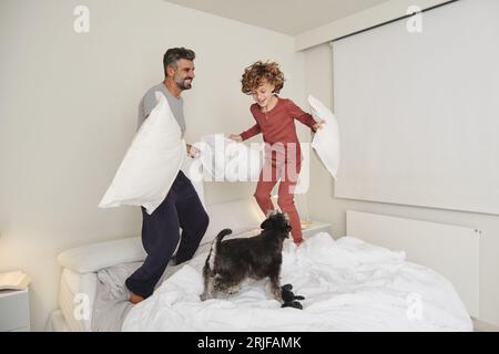 Corps entier de père adulte joyeux et fils aux cheveux bouclés en pyjama sautant sur le lit avec des oreillers tout en jouant avec le chien Banque D'Images