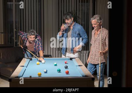 Amis masculins matures positifs dans des tenues décontractées debout près de la table de billard et jouer au billard tout en buvant des cocktails dans une salle moderne avec bras en cuir Banque D'Images