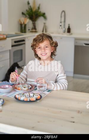 Enfant heureux avec les cheveux bouclés souriant et regardant la caméra assis à table et mangeant de savoureux rouleaux de sushi avec des baguettes roses dans la cuisine à la maison Banque D'Images