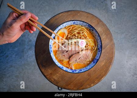Vue de dessus de la récolte méconnaissable personne tenant des baguettes tout en mangeant une délicieuse soupe de ramen japonaise avec des nouilles et de la viande de porc garnie d'oeuf et Banque D'Images