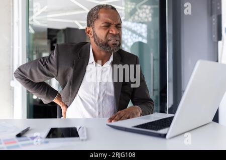 Homme d'affaires afro-américain mûr malade ayant mal au dos grave, homme en costume d'affaires au travail assis surtravaillé et surfatigué. Banque D'Images