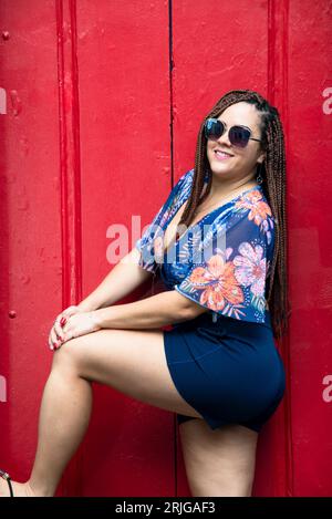 Jolie femme portant des lunettes de soleil avec des cheveux tressés debout dans une porte rouge. Pelourinho, Brésil. Banque D'Images