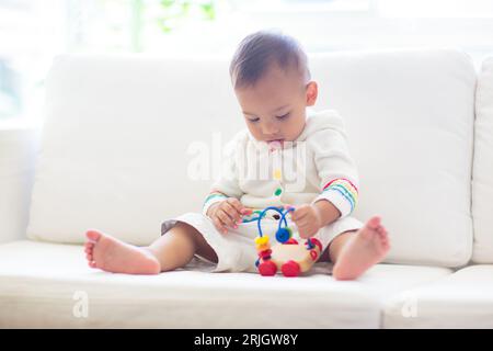 Bébé garçon asiatique jouant avec train de jouets coloré dans la chambre blanche ensoleillée. Bébé avec des jouets éducatifs. Enfant assis sur un canapé blanc. Développement précoce pour Banque D'Images