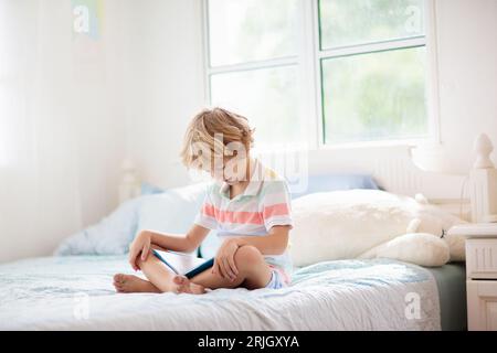 Les enfants lisent un livre dans le lit.Chambre pour enfant.Un petit garçon lit des livres sur un lit blanc avec une literie colorée.Enfants en pépinière.Enfant garçon jouant à l'intérieur. Banque D'Images