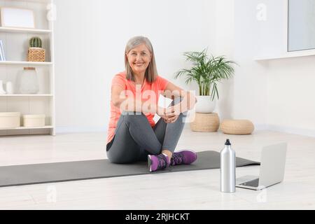 Femme âgée en vêtements de sport assis sur un tapis de fitness près d'un ordinateur portable à la maison Banque D'Images