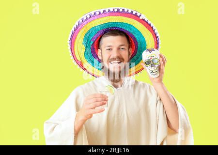 Beau jeune homme en sombrero mexicain avec crâne peint et plan de tequila sur fond jaune Banque D'Images