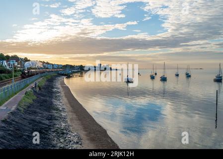 Chalkwell, Southend on Sea, Essex, Royaume-Uni. 23 août 2023. Un train à vapeur a quitté Southend on Sea tôt le matin en direction de Bristol Temple Meads, en passant le long de l'estuaire calme et plat de la Tamise à Chalkwell. Le train spécial organisé par la Railway Touring Company est transporté par l'ancienne locomotive à vapeur LMS Jubilee Class 45690 Leander. Leander est l'une des locomotives à vapeur autorisées à circuler sur les lignes principales pour des voyages spéciaux et a été construite en 1936. Il a été renuméroté 45690 par British Railways en 1948 à la suite de la nationalisation. Yachts sur l'eau de l'estuaire qui reflète la lumière de l'aube Banque D'Images