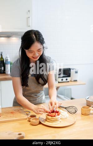 A beautiful young Asian woman adds fresh strawberries on top of her pancakes, enjoying preparing her fancy breakfast in the kitchen. Stock Photo