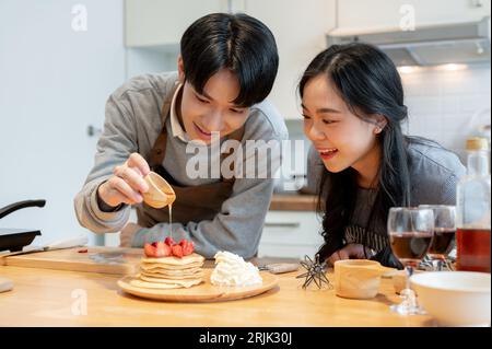 Un beau jeune homme asiatique verse du sirop sur des crêpes, aime cuisiner des crêpes avec sa petite amie dans la cuisine le week-end. Banque D'Images