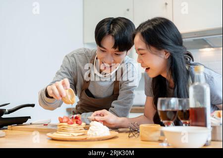 Un beau jeune homme asiatique verse du sirop ou du miel sur des crêpes, aime cuisiner des crêpes avec sa petite amie dans la cuisine et dépenser heureux, quali Banque D'Images