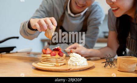 Close-up image of a handsome young Asian man is pouring syrup on pancakes, enjoys cooking pancakes with his girlfriend in the kitchen on the weekend. Stock Photo