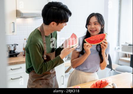 Un charmant couple asiatique aime manger de la pastèque ensemble dans la cuisine, passer du temps amusant ensemble à la maison. Date de cuisson, vie domestique romantique, amour Banque D'Images