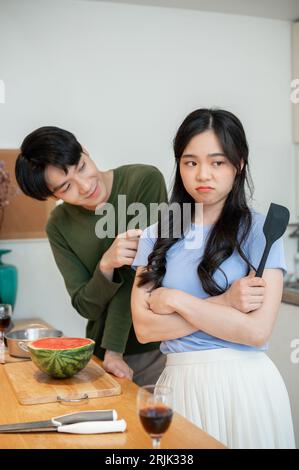 Un jeune petit ami asiatique mignon et collant demande pardon à sa petite amie en colère après s'être disputé tout en cuisinant dans la cuisine ensemble. Banque D'Images