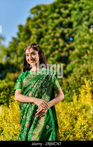 jolie et souriante femme indienne en sari regardant la caméra tout en posant près des plantes dans le parc Banque D'Images