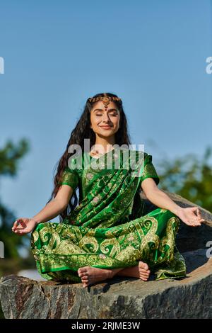 femme indienne souriante pieds nus en sari méditant sur la pierre avec ciel bleu sur fond Banque D'Images