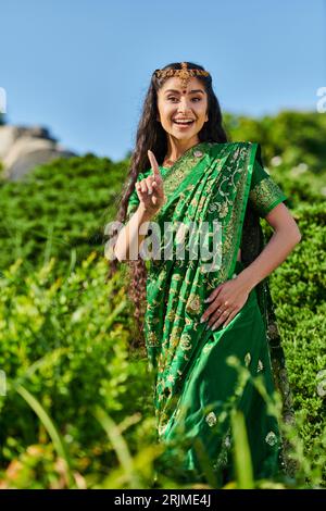 joyeuse jeune femme indienne dans sari pointant du doigt tout en se tenant près des plantes dans le parc Banque D'Images