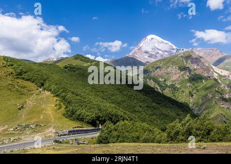 Le cône volcanique enneigé de Kazbek (5 047 m) domine la vallée boisée de Terek près de Stepantsminda, en Géorgie ; en contrebas, la route militaire géorgienne traverse la vallée. L'autoroute militaire géorgienne relie Tbilissi à la frontière russe via le district de Kazbegi et est l'une des plus anciennes routes de montagne du Caucase. Stepantsminda-Sameba, Gergeti, municipalité de Kazbegi, Mtskheta-Mtianeti, 4700, Géorgie Banque D'Images