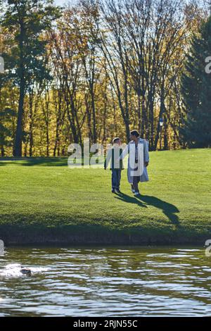 heureuse mère afro-américaine et fils marchant vers le lac, femme et garçon en vêtements d'extérieur, nature, automne Banque D'Images