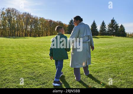 vue arrière d'une femme afro-américaine marchant avec son fils dans le parc, herbe verte, tenues d'automne, vêtements d'extérieur Banque D'Images