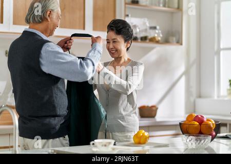 mari asiatique senior aimant aidant femme à mettre sur le tablier dans la cuisine à la maison Banque D'Images