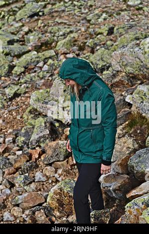 Portrait d'une femme souriante randonneuse dans une veste verte et un pantalon de voyage noir marche sur une pente rocheuse de montagne. Concept de voyage et de mode de vie actif. Banque D'Images