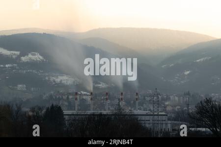 Fumée sortant des cheminées de l'installation de chauffage le soir, montagne Ponir avec le sommet éclairé par le soleil et le ciel incandescent en arrière-plan Banque D'Images