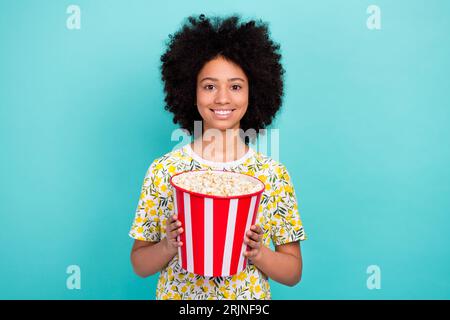 Photo de mignonne fille joyeuse porter des vêtements à la mode mains tiennent le panier délicieux pop corn isolé sur fond de couleur cyan Banque D'Images