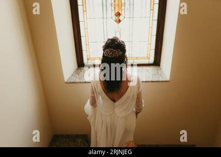 Une jeune femme portant une robe de mariée blanche se tient dos à la caméra, éclairée par la lumière qui circule à travers un vitrail Banque D'Images