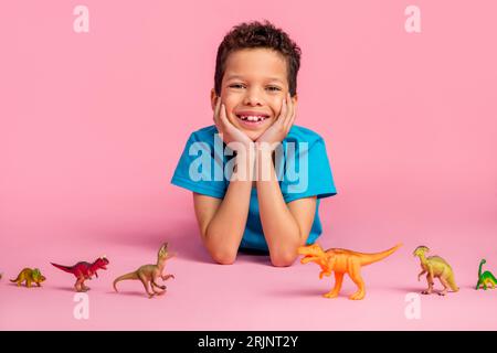 Photo de joyeux petit garçon doux habillé t-shirt bleu jouant des jouets de dinosaures en plastique isolé fond de couleur rose Banque D'Images