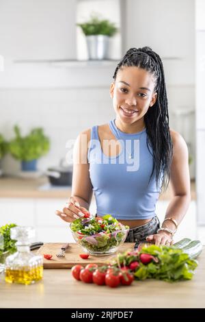 Jeune femme afro-américaine prépare une salade saine, ajoute des tomates. Banque D'Images