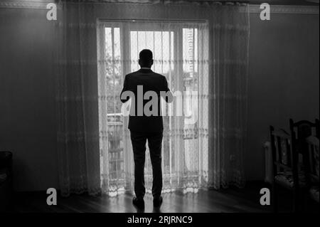 A man in a classic suit is standing by the window. A man in trousers and a jacket looks out the window Stock Photo