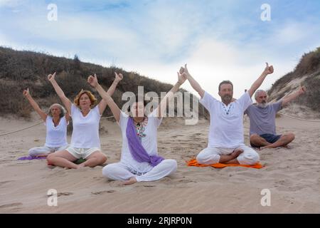 Groupe d’hommes et de femmes pratiquant la méditation et le yoga sur le sable de la plage, les bras levés, l’éradicateur de l’ego Banque D'Images