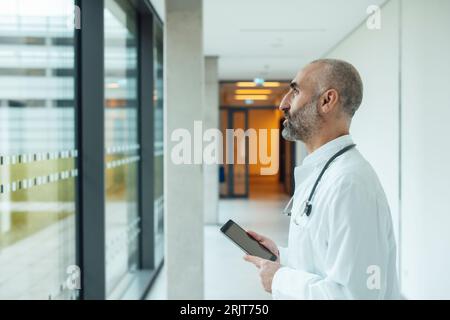 Médecin mûr réfléchi avec tablette PC regardant par la fenêtre à l'hôpital Banque D'Images