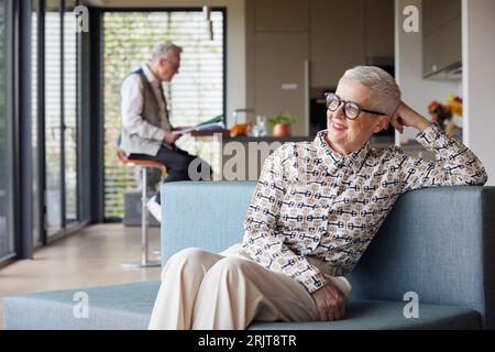 Femme âgée souriante assise sur le canapé à la maison avec l'homme en arrière-plan Banque D'Images