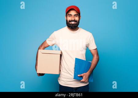 Réception de colis. Livreur indien tenant le presse-papiers et la boîte en carton, livrant des marchandises et de la nourriture, fond bleu Banque D'Images