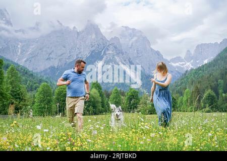 Homme et femme courant avec chien sur l'herbe devant les montagnes Banque D'Images