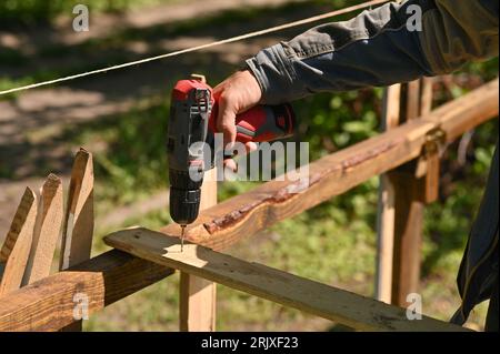 Un homme perce un trou dans une planche de bois avec une perceuse. en extérieur. Banque D'Images