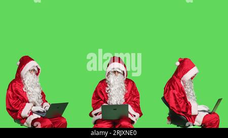 père noël moderne travaille sur ordinateur portable, fond greenscreen dans le studio. Père noël assis sur une chaise et utilisant un pc sans fil pour la navigation sur le site Web en ligne, la recherche d'adresses d'enfants. Banque D'Images