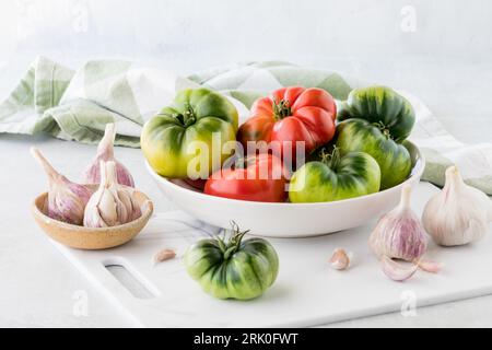 Un bol plein de tomates héritées fraîchement cueillies, sur une planche à découper blanche. Banque D'Images