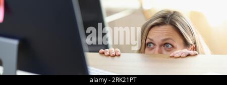 Une femme de direction effrayée cachée derrière le bureau et regardant l'écran de l'ordinateur. Banque D'Images