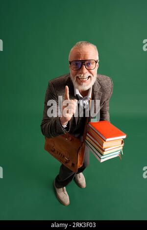 Portrait en longueur de l'homme âgé avec barbe, professeur portant costume vintage avec grande pile de livres et sac d'affaires brun sur fond vert. Banque D'Images