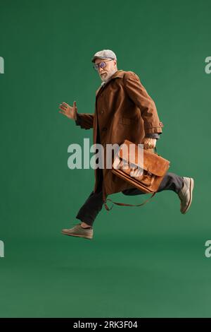 Portrait en longueur de l'homme senior en cours d'exécution avec barbe, détective portant costume vintage hatand avec grand sac d'affaires sur fond vert. Banque D'Images