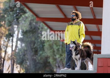 Un homme hispanique portant une veste jaune et des lunettes de soleil tenant la laisse de son chien Malamute d'Alaska Banque D'Images