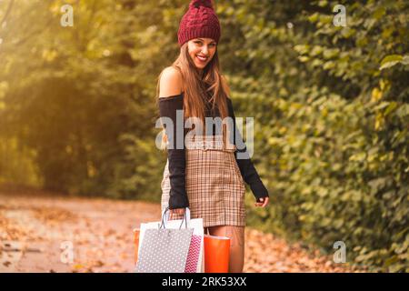A young female wearing a plaid skirt and a stylish hat is walking and smiling while carrying shopping bags Banque D'Images